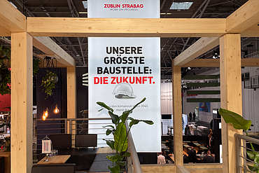 Messestand mit Holzrahmen und Pflanzen. Im Zentrum ein Banner mit den Logos von ZÜBLIN und STRABAG sowie dem Slogan ‚WORK ON PROGRESS‘. Darunter der Text: ‚Unsere größte Baustelle: Die Zukunft. Wir werden klimaneutral bis 2040. Hand drauf!
