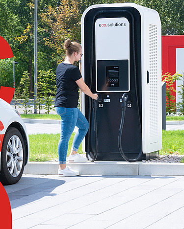 Picture of an e-car being charged by a woman at an e-charging station using clean electromobility. Next to it are large red displays of C, O and 2.