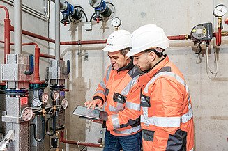 Foto von zwei Personen in Schutzkleidung, die vor einer technischen Anlage auf ein Tablet schauen
