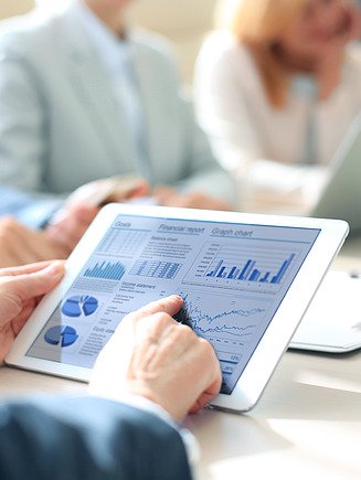 Image of a person holding a tablet with business graphics and statistics while several people in business attire sit at a meeting table in the background.