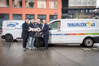 STRABAG PFS Triburuzek Foto von vier Männern, die sich vor einem Gebäude und zwei Autos einen großen Schlüssel übergeben