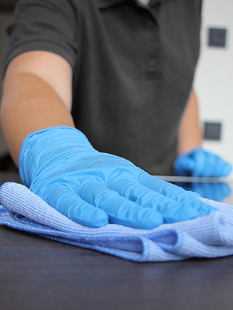 Bild von einer Hand mit blauem Handschuh, welche mit einem blauen Tuch eine Oberfläche reinigt