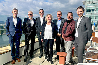 Foto von sieben Menschen, die nebeneinander auf einem Dach stehen bei Sonnenscheinund in die Kamera lächeln