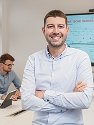 Mann mit verschränkten Armen steht im Vordergrund, im Hintergrund andere Personen und ein Präsentationsbildschirm