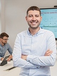 Mann mit verschränkten Armen steht im Vordergrund, im Hintergrund andere Personen und ein Präsentationsbildschirm