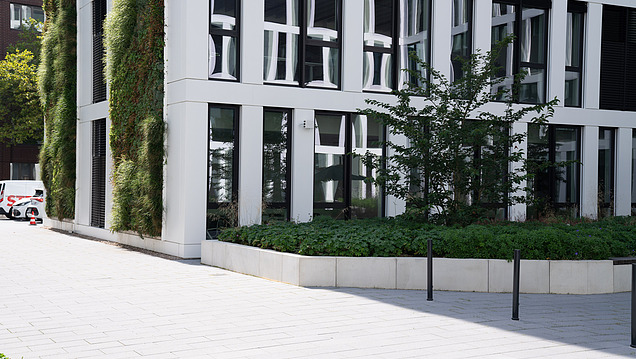 Moderní fasáda budovy s vertikální zelení a osázeným venkovním prostorem ve slunném městském prostředí.