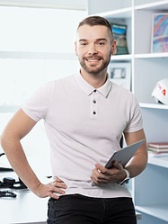 Person mit Tablet lehnt sich in modernem Büro an einen Tisch, Regale im Hintergrund.