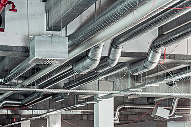 Mehrere Lüftungsrohre und Kabel verlaufen entlang einer industriellen Decke in einem Gebäude.