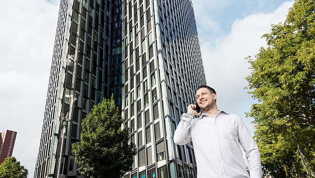 Usměvavý muž v košili telefonuje před prosklenou kancelářskou věží se stromy.