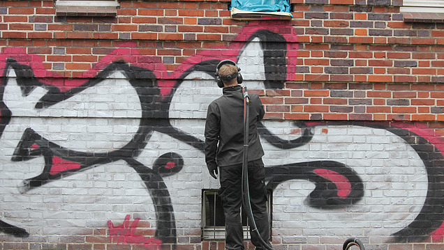 Eine Person in Schutzkleidung steht vor einer mit Graffiti besprühten Mauer und reinigt diese