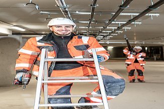 Muž v ochranném oděvu stojí na žebříku a usmívá se; v pozadí je muž, který nese dlouhý stavební prvek