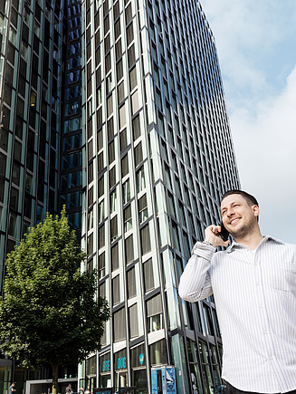 Eine Person telefoniert vor einem modernen Bürogebäude mit Bäumen und weiteren Gebäudeteilen im Hintergrund.