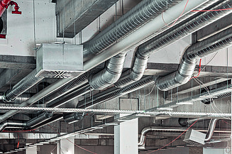 Mehrere Lüftungsrohre und Kabel verlaufen entlang einer industriellen Decke in einem Gebäude.