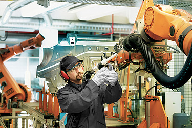 Ein Arbeiter in Schutzkleidung bedient einen großen orangefarbenen Roboterarm in einer automatisierten Fertigungshalle.