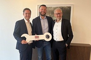 Schlüsselübergabe B2 Assets Foto von drei Männern, die nebeneinander stehen und sich einen großen Schlüssel überreichen