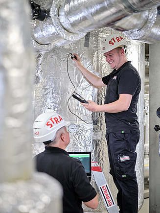Zwei STRABAG-Mitarbeiter arbeiten mit Messgeräten an isolierten Rohrleitungen in einer Industrieumgebung.