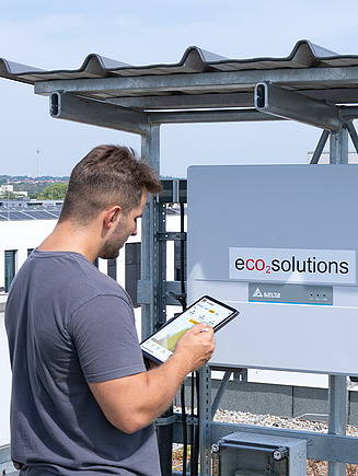 Obrázek zaměstnance společnosti STRABAG PFS, který pomocí tabletu kontroluje fotovoltaický systém na střeše a stojí před technickým systémem s nálepkou eco2solutions.