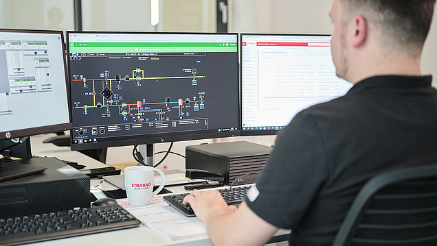 Ein Mann arbeitet an einem Schreibtisch mit drei Monitoren, die technische Diagramme und Tabellen anzeigen