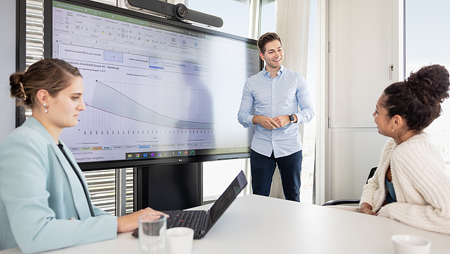 Vorbereitung Regelbetrieb STRABAG PFS Österreich Ein Mann präsentiert ein Diagramm auf einem großen Bildschirm, während zwei Frauen am Tisch zuhören
