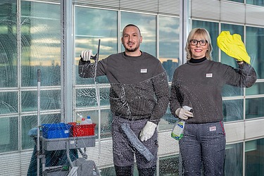 Zwei Personen reinigen die Fenster eines Gebäudes mit Reinigungswerkzeugen und -mitteln.t.“