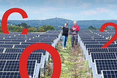 Bild von einer PV-Anlage auf einem begrünten Dach und zwei Menschen im Hintergrund. Auf dem Dach stehen große rote Aufsteller von C, O und 2.