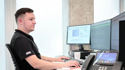 Muž v košili u stolu se třemi obrazovkami, telefon s displejem na pravém okraji obrazu.