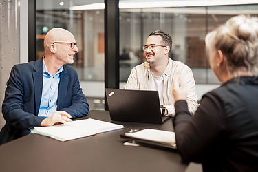 Tři kolegové na schůzce, dva muži hovoří na notebooku, žena zezadu, moderní kancelářské prostředí.