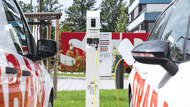 Bild von zwei E-Autos mit STRABAG Logo, welche an eine E-Ladesäule angeschlossen sind und aufgeladen werden.