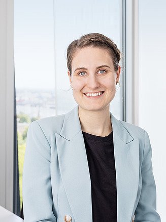 Person in hellblauem Blazer und schwarzem Oberteil steht vor einem Fenster mit unscharfer Stadtlandschaft im Hintergrund