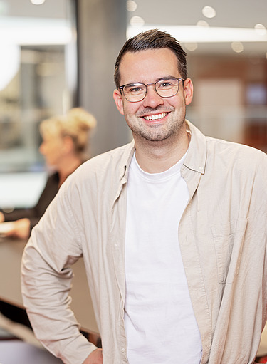 Photo of a man standing in front of a desk where 2 people are working
