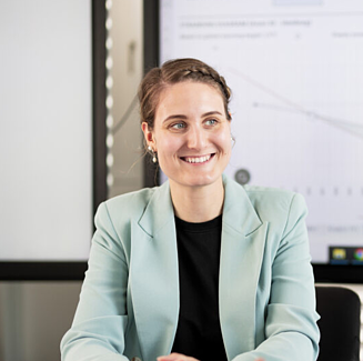 Person in hellblauem Blazer und schwarzem Oberteil, im Hintergrund verschwommen ein Präsenationsbildschirm