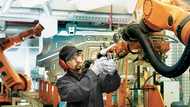 Ein Arbeiter in Schutzkleidung bedient einen großen orangefarbenen Roboterarm in einer automatisierten Fertigungshalle.