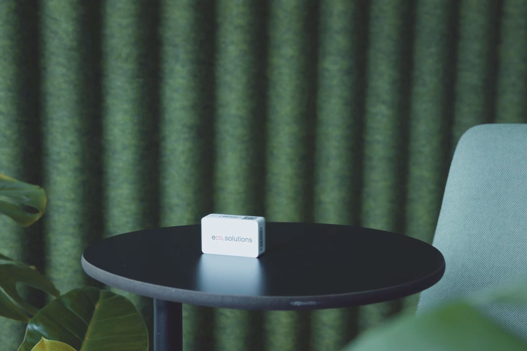 Bild von einem kleinen eco2state Sensor, welcher auf einem runden Tisch vor grünem Hintergrund steht.