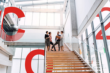 Picture of two people walking in front of a glass office building. Next to them are large red displays of C, O and 2.