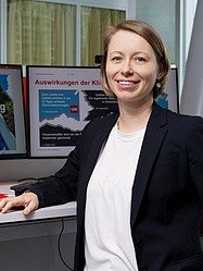 Frau steht in einem Büro vor zwei Monitoren mit deutschsprachigem Text.