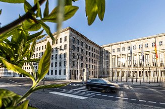 Bild von einem historischen Gebäude, die Vorfahrt ist beflaggt, auf der Straße ist ein vorbeifahrendes Auto zu sehen
