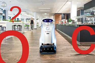 Bild von einem Reinigungsroboter in einer Kantine. Weiterhin sind große rote Aufsteller von C, O und 2 zu sehen.