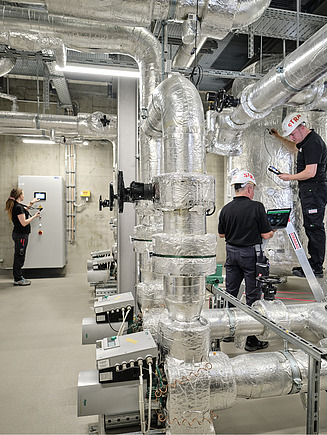 Photo of a boiler room with silver pipes in which three people are working