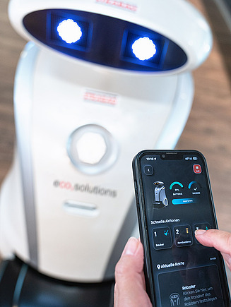 Bild von einem mit einer Hand gehaltenen Smartphone, welches eine Anwendung zeigt und im Hintergrund befindet sich ein Reinigungsroboter