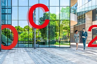 Photo of a glass building with two people passing by and big red letters O, C, 2