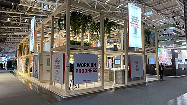 STRABAG Messestand mit Holzrahmen und Innovationsbotschaften Messestand mit offener Holzrahmenstruktur, hängenden Pflanzen und mehreren Postern mit Slogans wie ‚WORK ON PROGRESS‘. Im Hintergrund weitere Messestände.