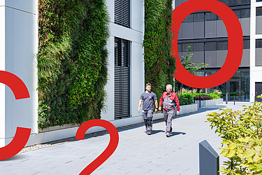 Image of two men walking alongside a vertically greened office building, in front of which are large red displays of C, O and 2.