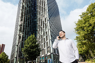 Eine Person telefoniert vor einem modernen Bürogebäude mit Bäumen und weiteren Gebäudeteilen im Hintergrund.