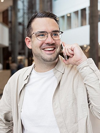 Person in weißem T-Shirt und hellbeiger Jacke steht im inneren eines großen Gebäudes und telefoniert