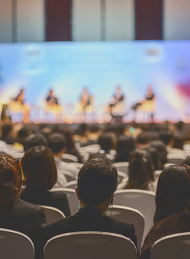 Picture of a conference situation, people are sitting in the foreground and people on a stage can be seen blurred in the background