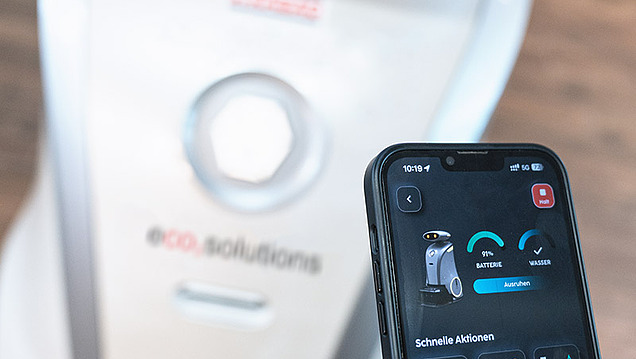 Bild von einem mit einer Hand gehaltenen Smartphone, welches eine Anwendung zeigt und im Hintergrund befindet sich ein Reinigungsroboter