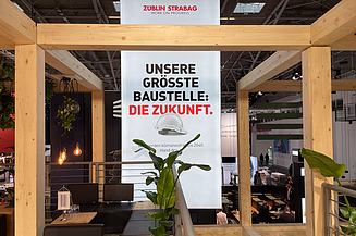 Messestand mit Holzrahmen und Pflanzen. Im Zentrum ein Banner mit den Logos von ZÜBLIN und STRABAG sowie dem Slogan ‚WORK ON PROGRESS‘. Darunter der Text: ‚Unsere größte Baustelle: Die Zukunft. Wir werden klimaneutral bis 2040. Hand drauf!
