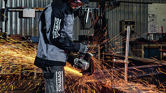 Eine Person in STRABAG-Schutzkleidung schleift Metall mit einem Winkelschleifer, während Funken sprühen.