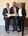 A photo of three men standing side by side, handing over a large key.