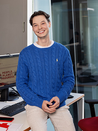 Eine Person sitzt lächelnd in einem modernen Büro am Schreibtisch mit zwei Monitoren, während eine STRABAG-Tasse und Büromaterialien sichtbar sind